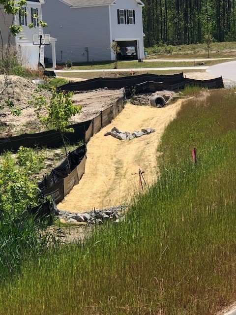 A newly constructed drainage ditch lined with straw and rocks runs beside a house and a street. Black silt fencing borders the ditch, and green grass grows in the surrounding area.